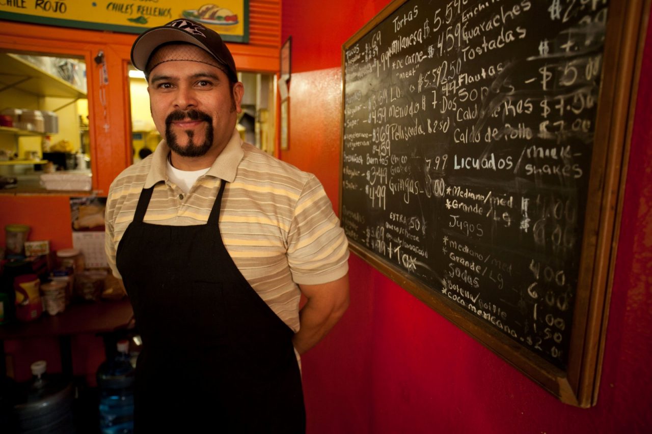 A chef standing by a restaurant menu printed on a chalkboard.