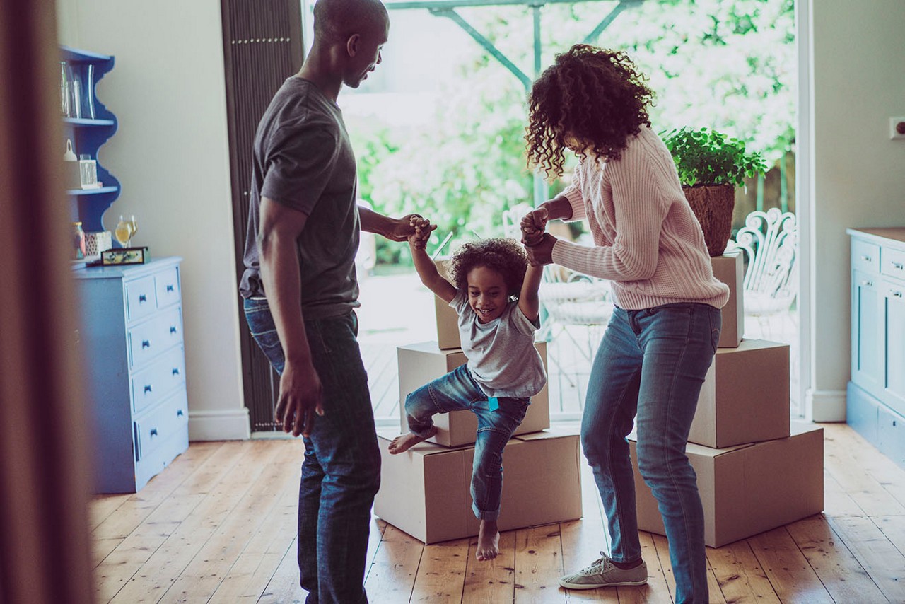 A photo of playful parents holding sons's hands in new house. Happy and playful family are with cardboard boxes. They are in casuals.