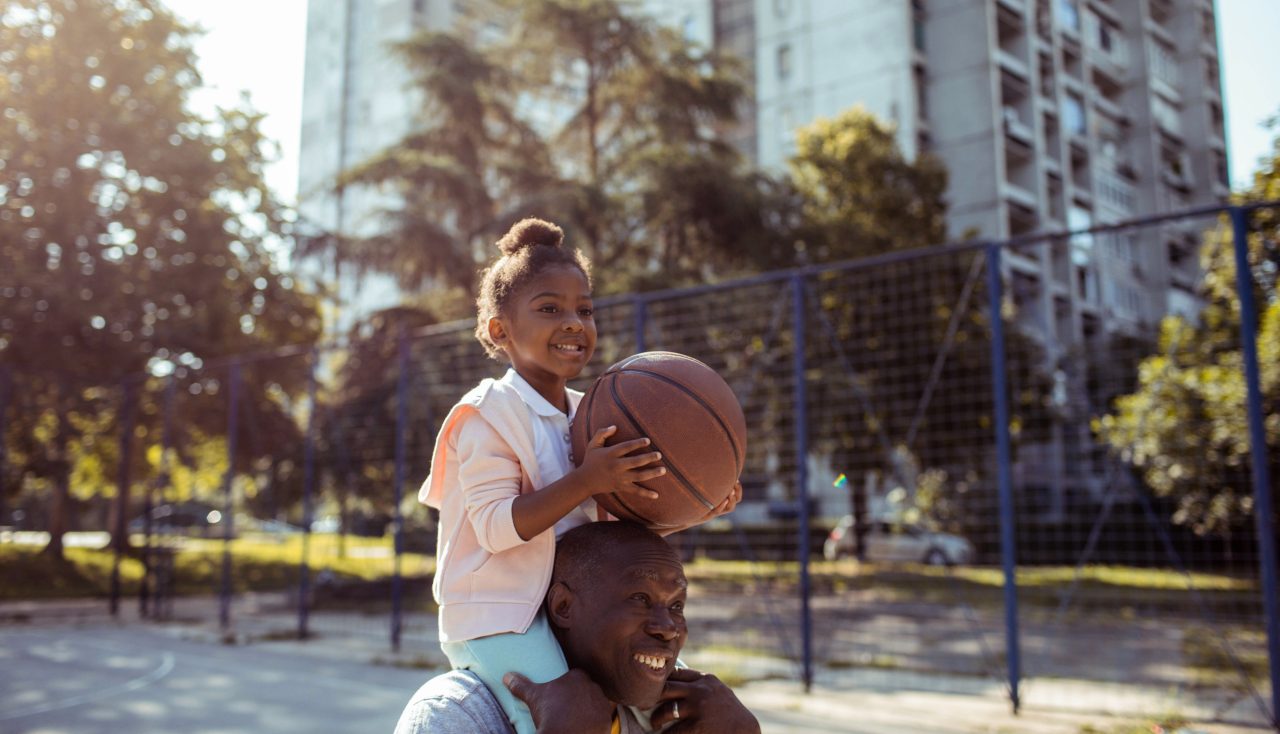 A young girl sitting on a man's shoulders while holding a basketball.