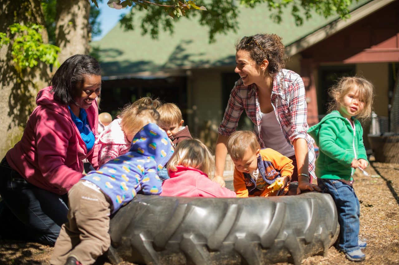 A daycare teacher playing with a group of young children outside.