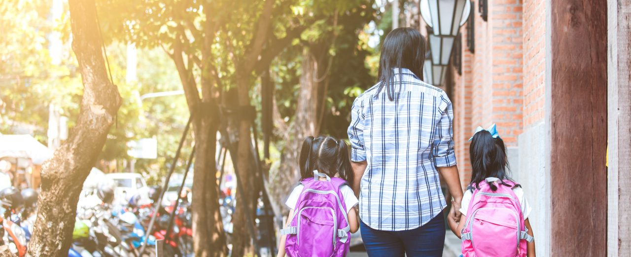 A mother and two young daughters with backpacks hold hands while walking on a sidewalk.